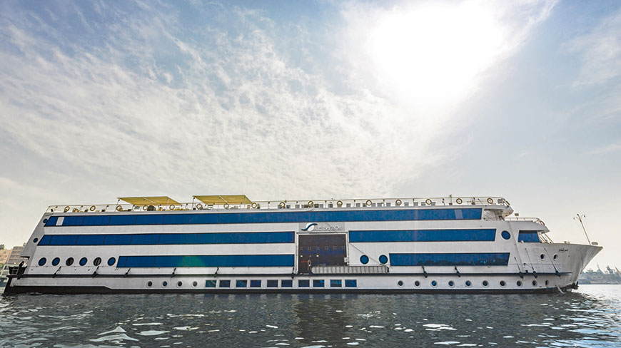 A large white and blue multi-decked river cruise ship sails on the water under a bright, cloudy sky.