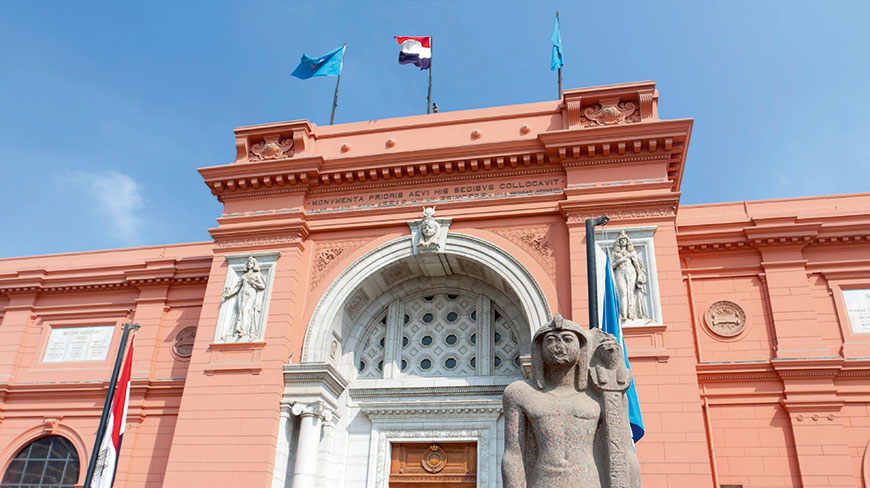 The grand, salmon-colored facade of the Egyptian Museum, with a large pharaoh statue standing before the arched entrance under a clear blue sky.