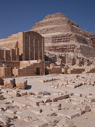 The Step Pyramid of Djoser towers over the stone ruins of the Saqqara necropolis under a clear blue sky.