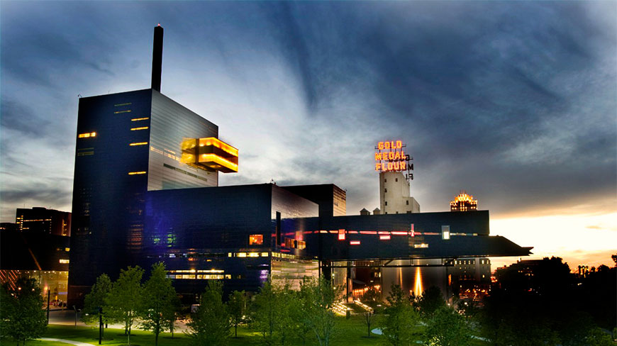 A twilight view of the modern Guthrie Theater in Minneapolis featuring illuminated windows and the iconic Gold Medal Flour sign in the background.
