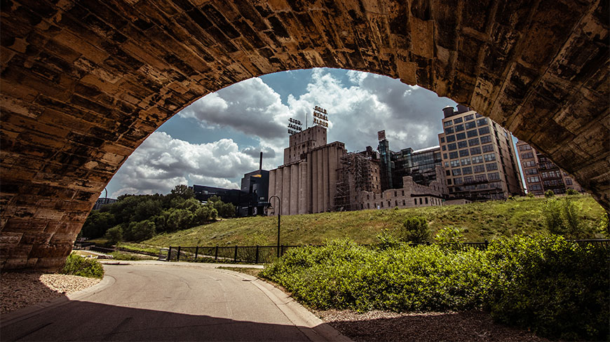 View of the Minneapolis Mill District and Gold Medal Flour sign framed by a large stone arch under a cloudy sky.