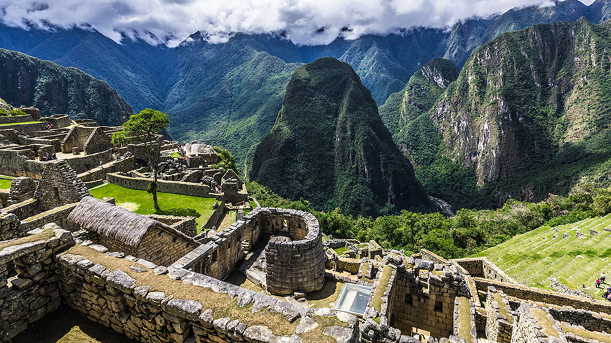 The ancient stone ruins of Machu Picchu sit on a green mountain ridge with steep, cloud-covered peaks in the background.