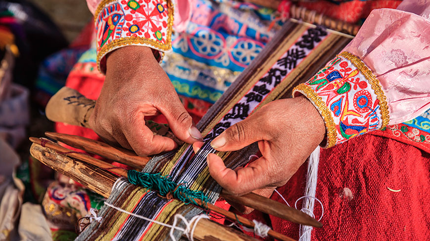 A close-up of a weaver's hands in colorful, embroidered sleeves creating a patterned textile on a traditional wooden loom.