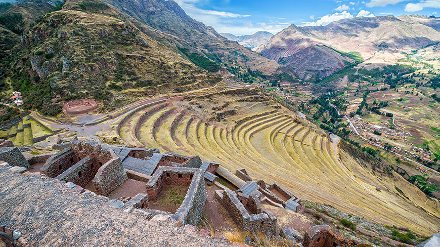 Ancient stone ruins overlook vast agricultural terraces carved into a steep mountainside under a blue sky with scattered clouds.