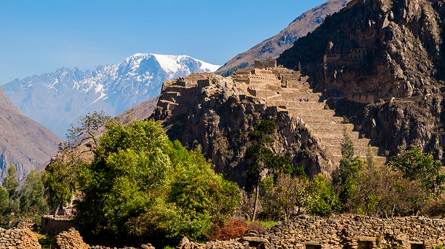 The stone terraces of the Ollantaytambo ruins climb a steep mountainside, with a snow-capped mountain peak rising in the background.