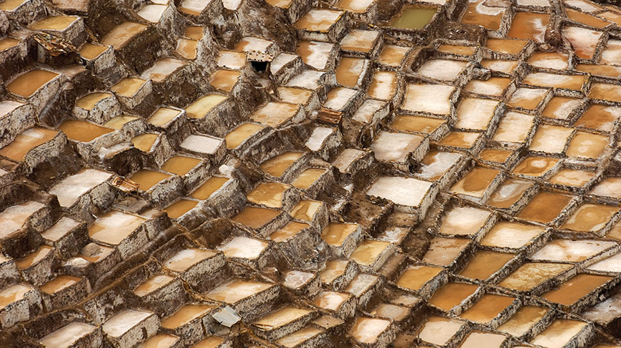 An overhead view shows a sprawling network of rectangular, earth-toned salt evaporation ponds terraced into a hillside.