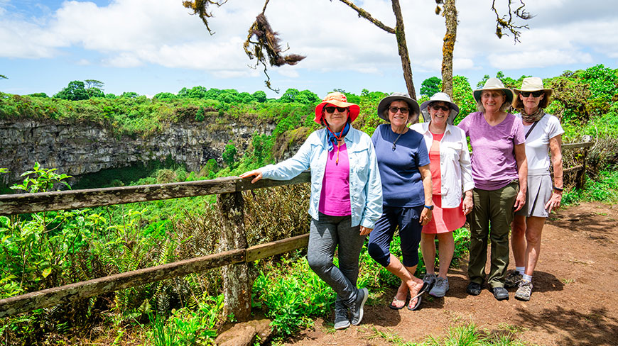 Five women in hats and sunglasses smile while posing for a photo by a wooden fence overlooking a lush green crater in the Galapagos.