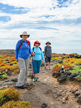Three women wearing hats and hiking gear smile while standing on a coastal trail in the Galapagos Islands.