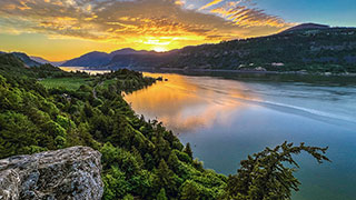 A vibrant sunset over the Columbia River Gorge in Oregon, with golden light reflecting on the water and lush green hillsides.