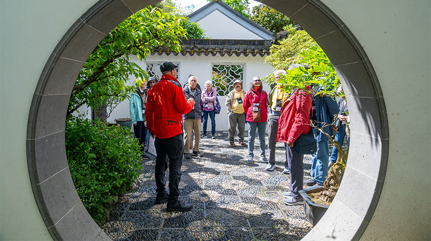 A tour group listens to a guide in a red jacket at the Lan Su Chinese Garden in Portland, viewed through a moon gate.