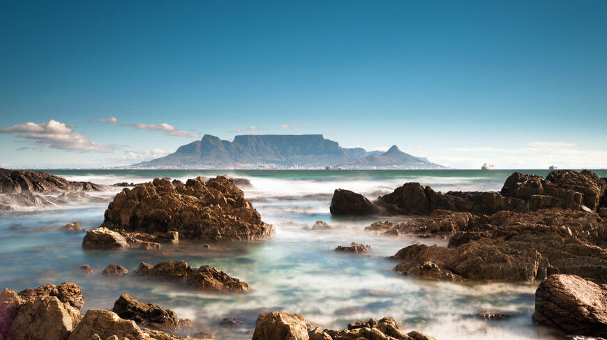 Table Mountain rises across a rocky coastline with misty ocean water in Cape Town, South Africa, under a clear blue sky.