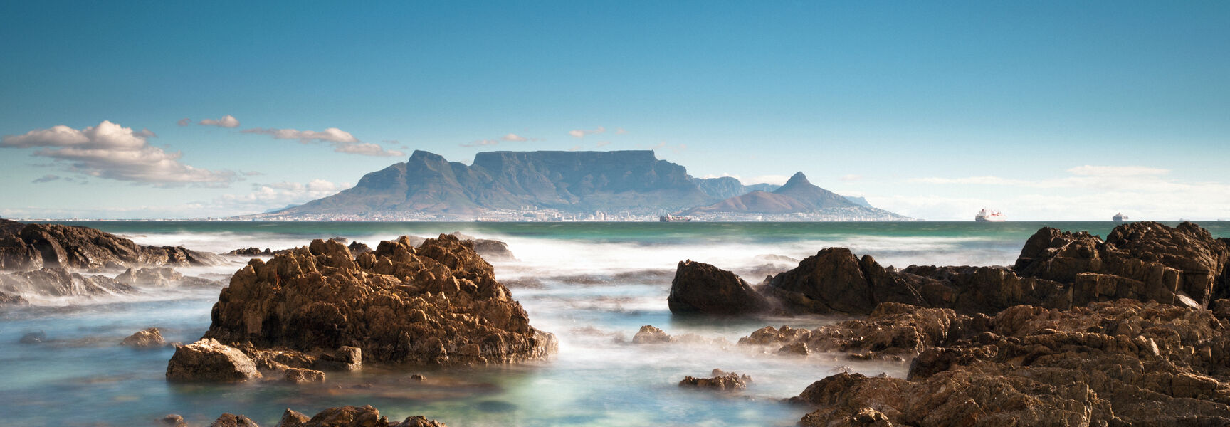 Table Mountain rises across a rocky coastline with misty ocean water in Cape Town, South Africa, under a clear blue sky.