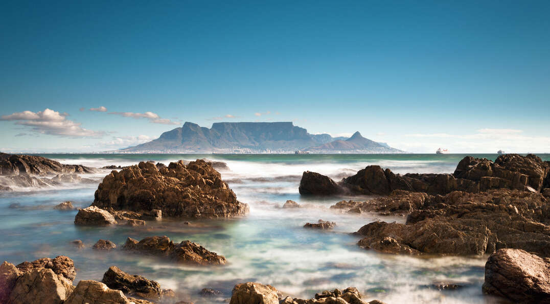 Table Mountain rises across a rocky coastline with misty ocean water in Cape Town, South Africa, under a clear blue sky.