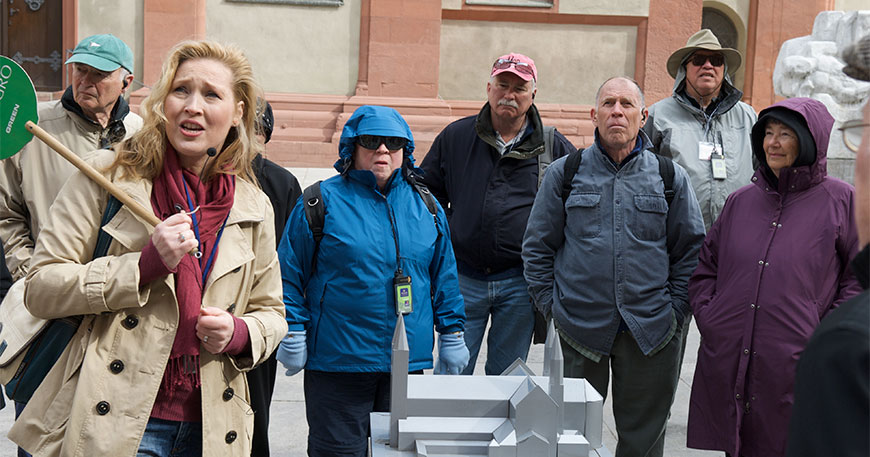A tour guide with a headset and green sign speaks to a group of tourists gathered around an architectural model outdoors.