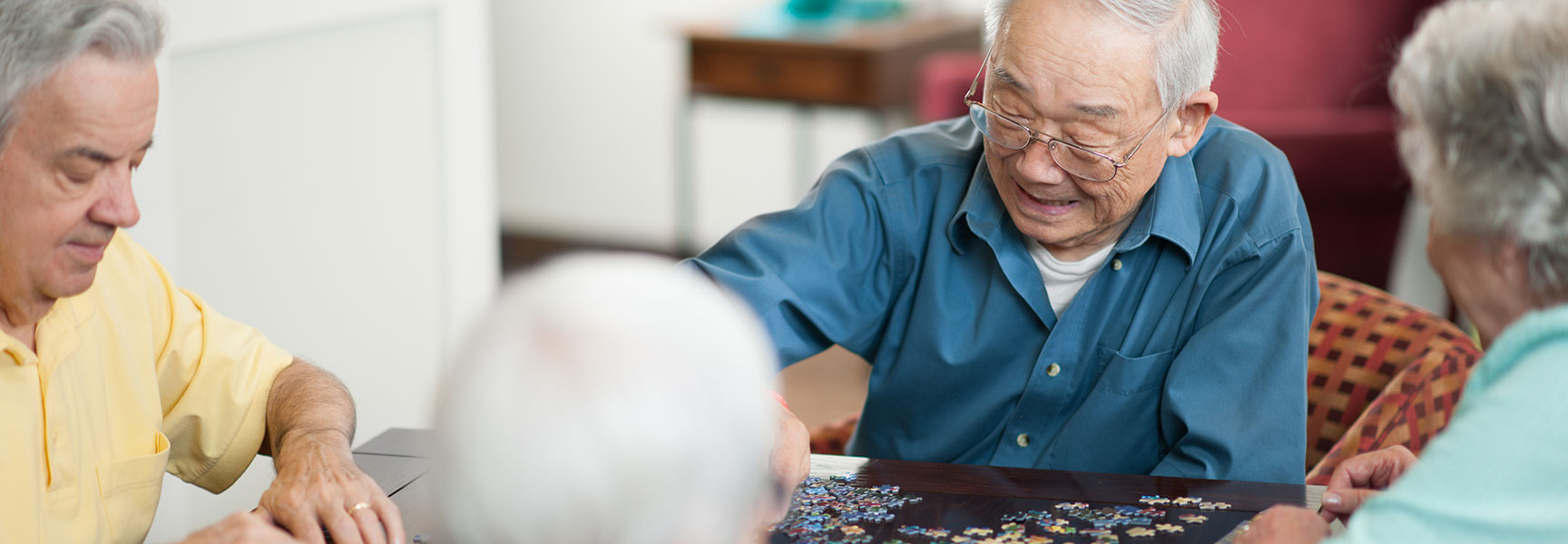 A group of seniors work together on a jigsaw puzzle at a table in a brightly lit room.
