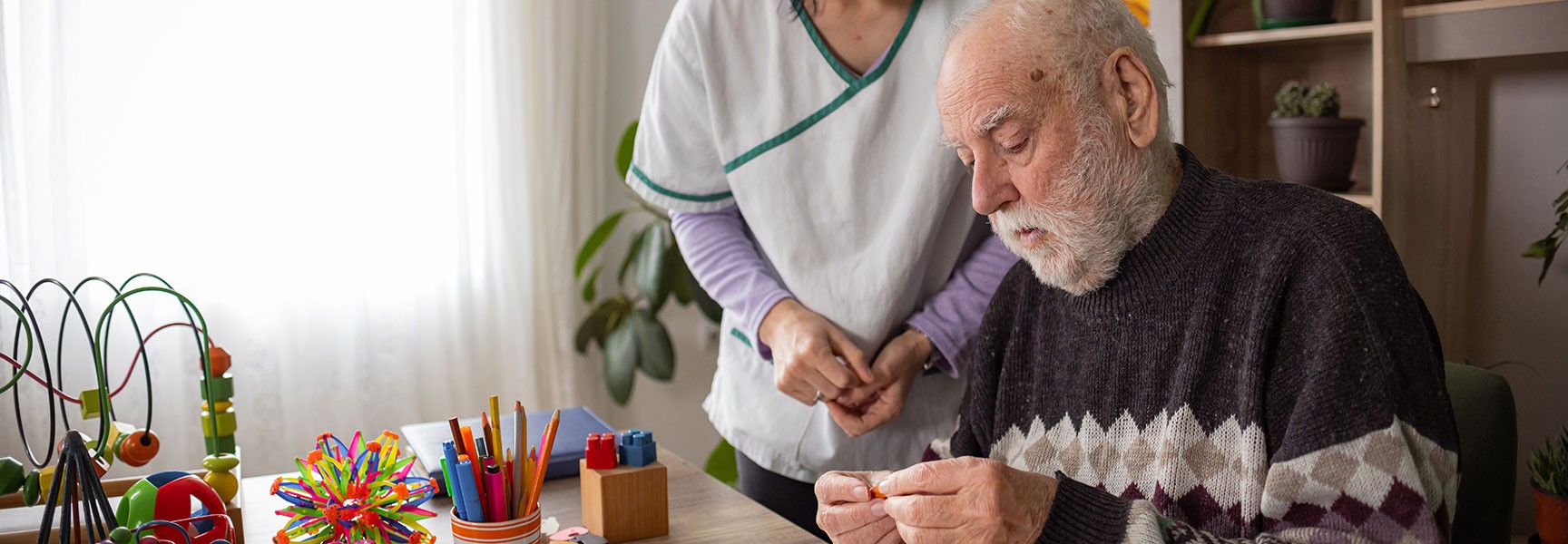 An older man with a white beard sits at a table doing a therapeutic activity while a caregiver stands behind him.