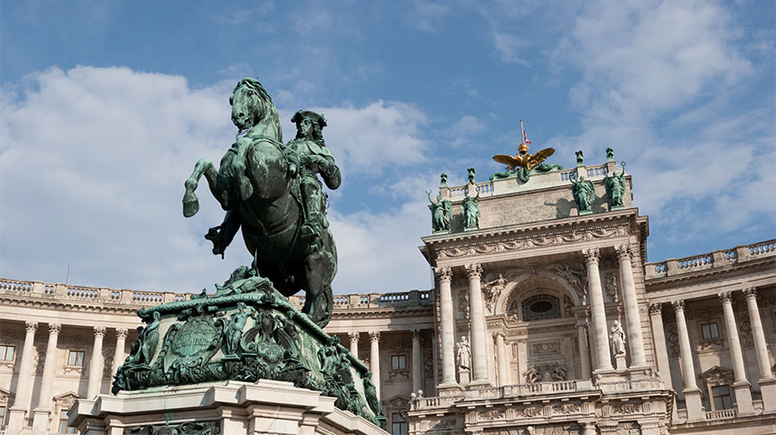 An equestrian statue stands in front of the ornate facade of the Vienna Hofburg Palace complex.