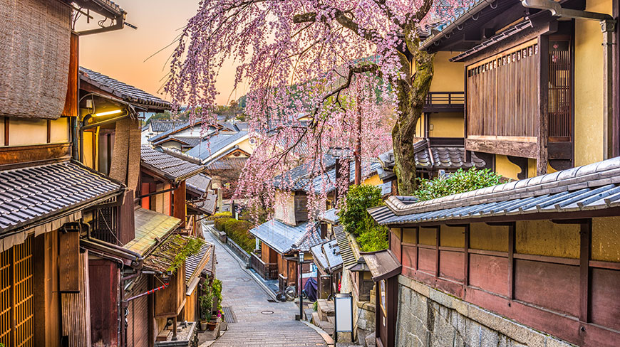 A narrow street in Kyoto's Higashiyama District with traditional houses and a blooming cherry blossom tree at sunset.