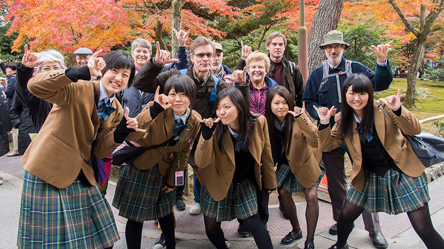 A group of Japanese schoolgirls in uniform and tourists pose for a photo outdoors with colorful autumn trees in the background.