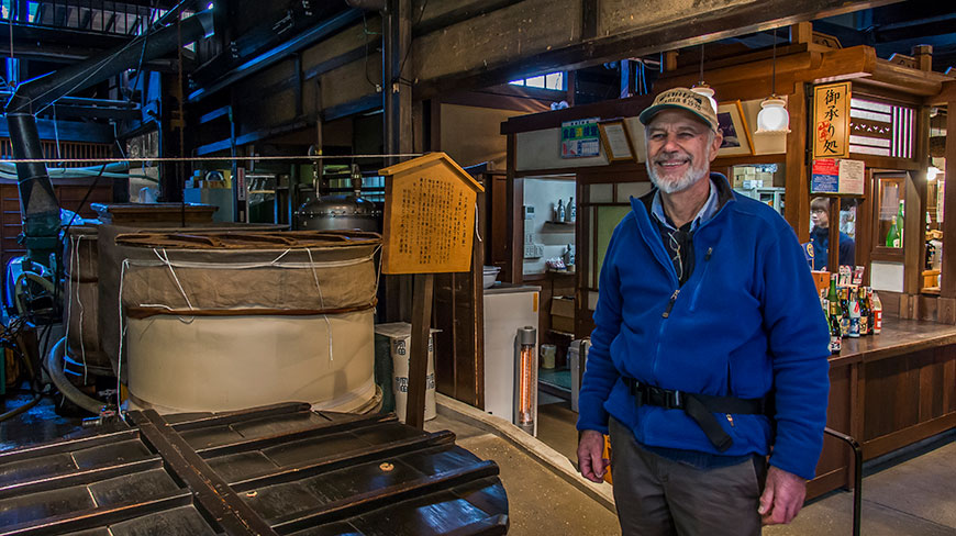 A smiling man in a blue fleece jacket and a cap stands inside a traditional Japanese sake brewery.