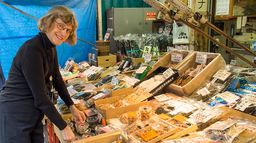 A woman in a black turtleneck sweater smiles while browsing a stall filled with packaged goods at a market in Tokyo.