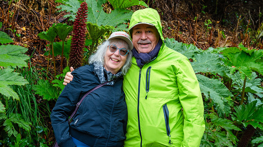 A smiling couple stands together in front of lush green foliage near the German Museum in Frutillar, Puerto Varas.