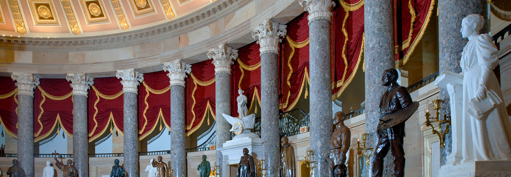 National Statuary Hall with marble columns, statues of historical figures, and ornate red curtains.