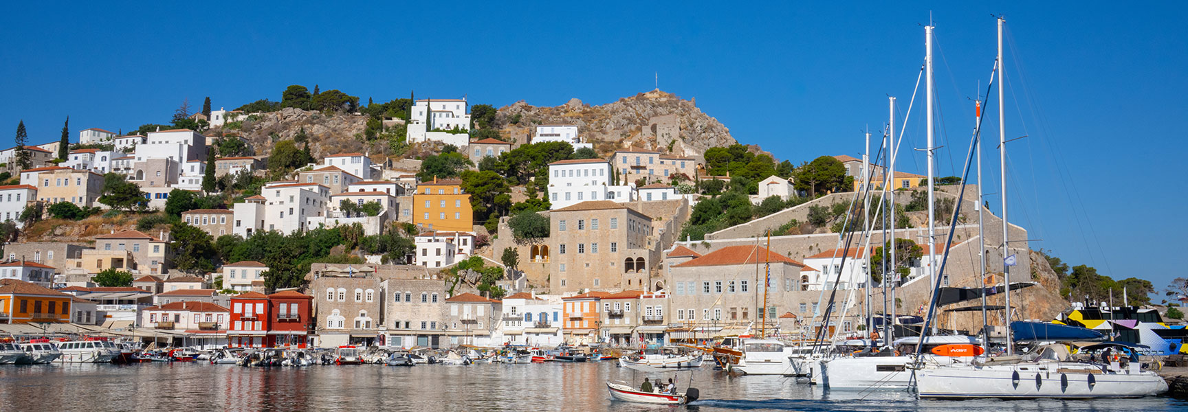 The sunny harbor of Hydra, Greece, is filled with boats in front of a hillside town of white and stone buildings under a clear sky.