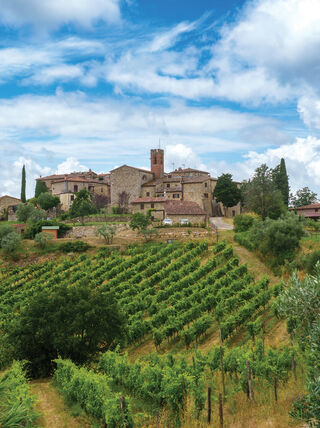 A stone village sits on a hilltop above sloping green vineyards under a vast blue sky filled with white clouds.
