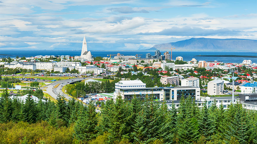 An elevated view of the Reykjavik cityscape featuring the Hallgrimskirkja church tower, coastal buildings, and distant mountains under a cloudy blue sky.