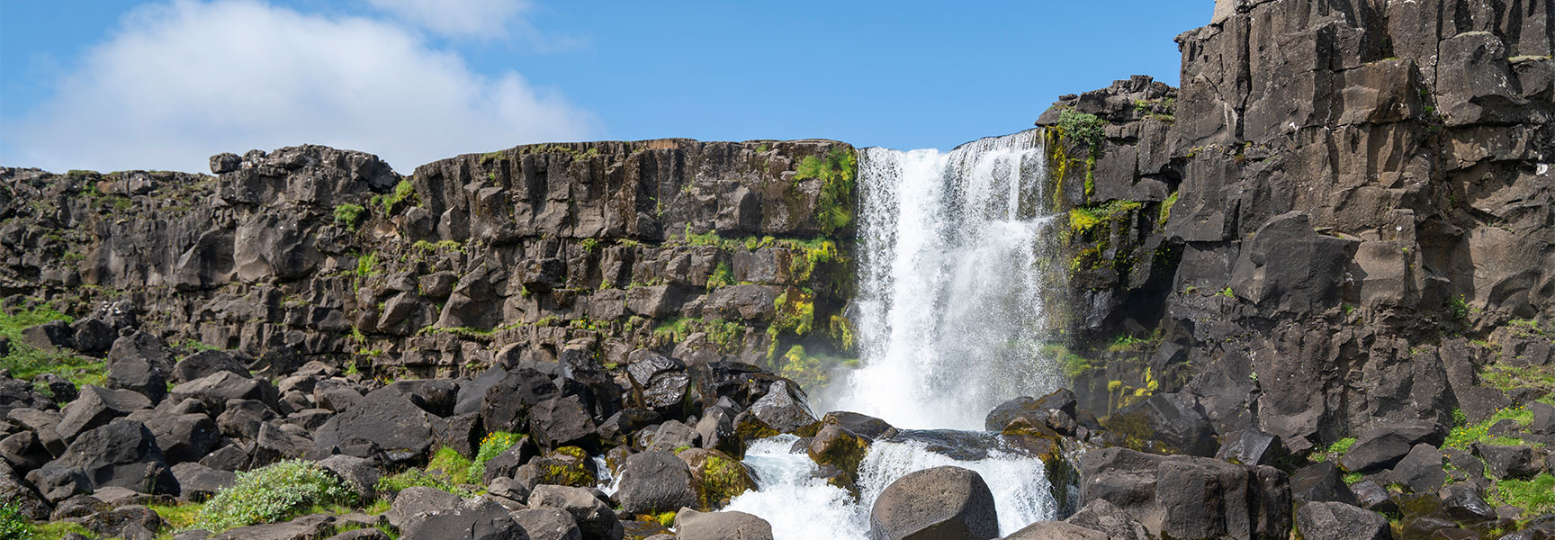 Oxararfoss waterfall flows over dark rock formations in Thingvellir National Park under a blue sky.