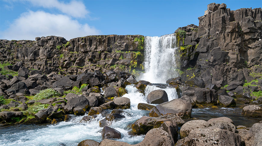 Oxararfoss waterfall flows over dark rock formations in Thingvellir National Park under a blue sky.