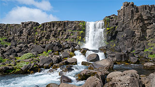 Oxararfoss waterfall flows over dark rock formations in Thingvellir National Park under a blue sky.