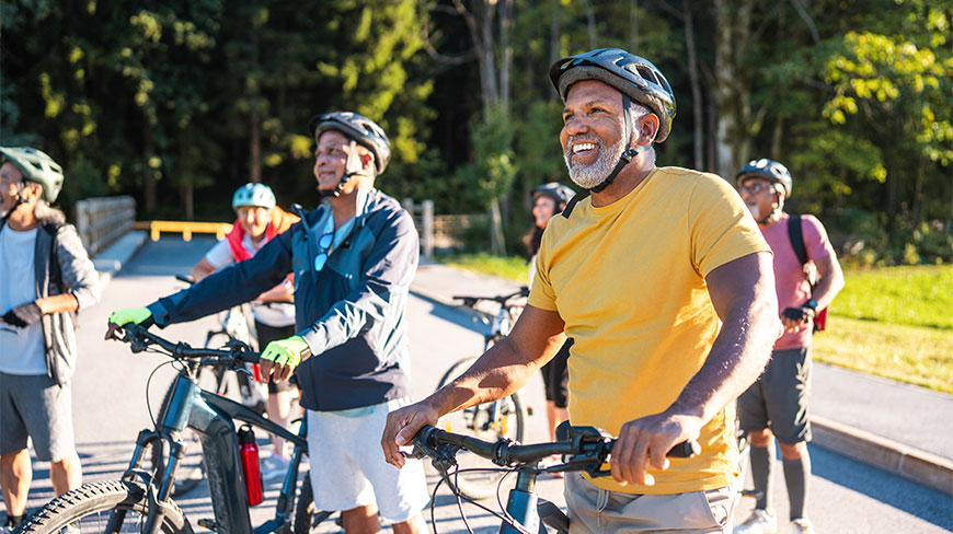 A diverse group of smiling older adults wearing helmets and athletic gear stand with their bicycles on a paved trail outdoors.