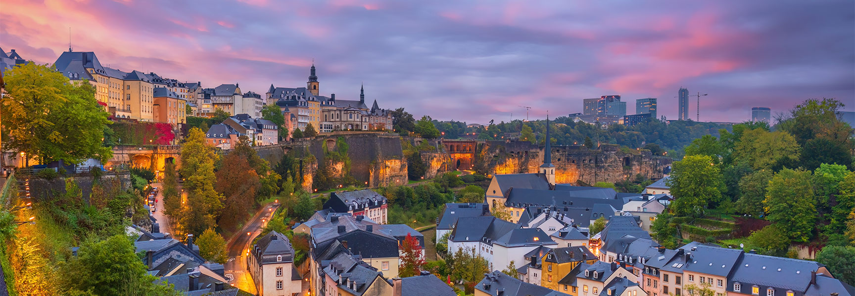 A panoramic view of the Luxembourg City old town at twilight with glowing streetlights and a colorful sunset sky.