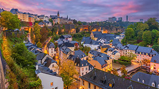 A panoramic view of the Luxembourg City old town at twilight with glowing streetlights and a colorful sunset sky.