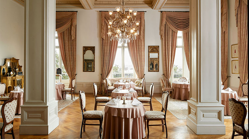 The elegant Pasa Salonu at the Pera Palace Hotel in Istanbul features a grand chandelier, tall pillars, and tables set for dining.