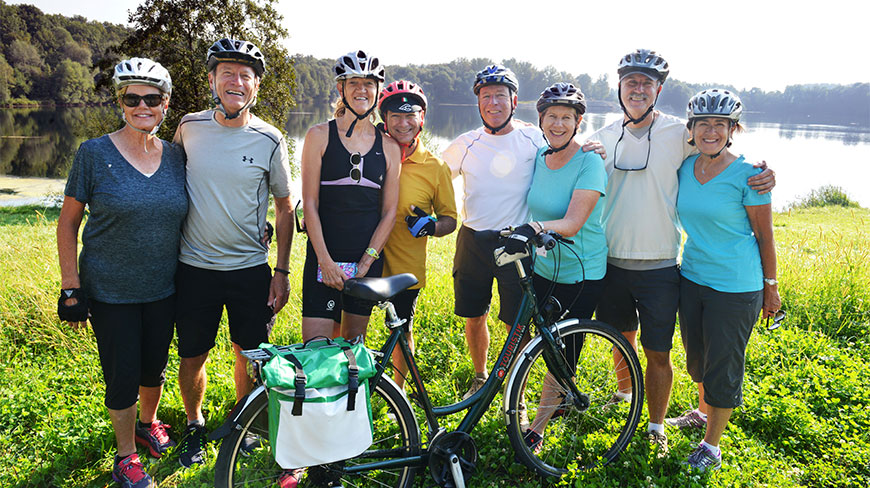 A group of eight smiling cyclists wearing helmets posing together outdoors with a touring bike in front of a calm lake.