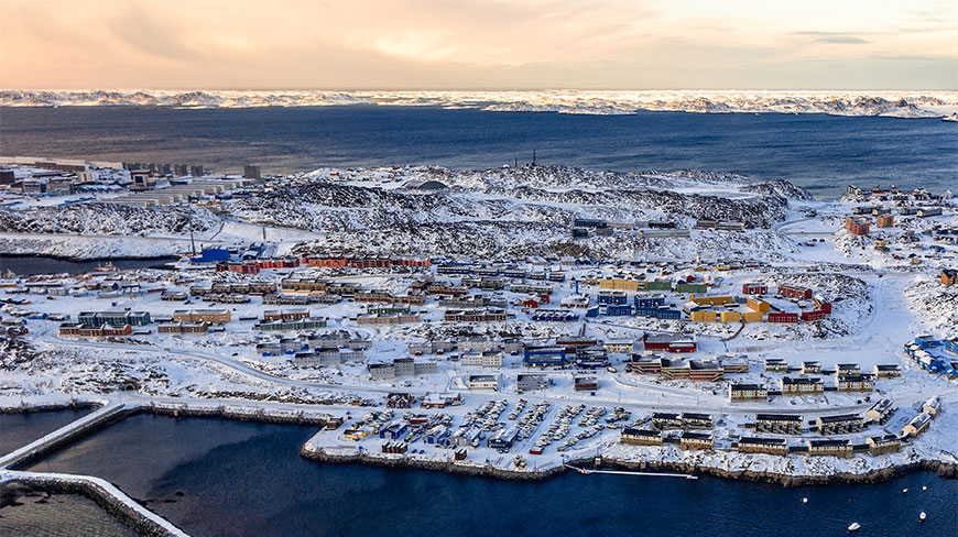 Aerial view of Nuuk, Greenland, featuring colorful buildings nestled in a snow-covered landscape along the coastline under a soft evening sky.
