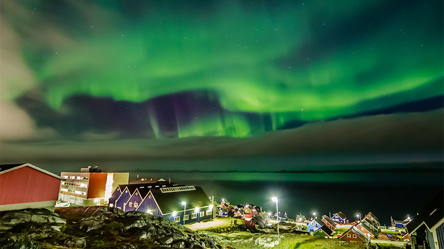 Vibrant green Northern Lights dance in the night sky over the colorful houses and coastal waters of Nuuk, Greenland.