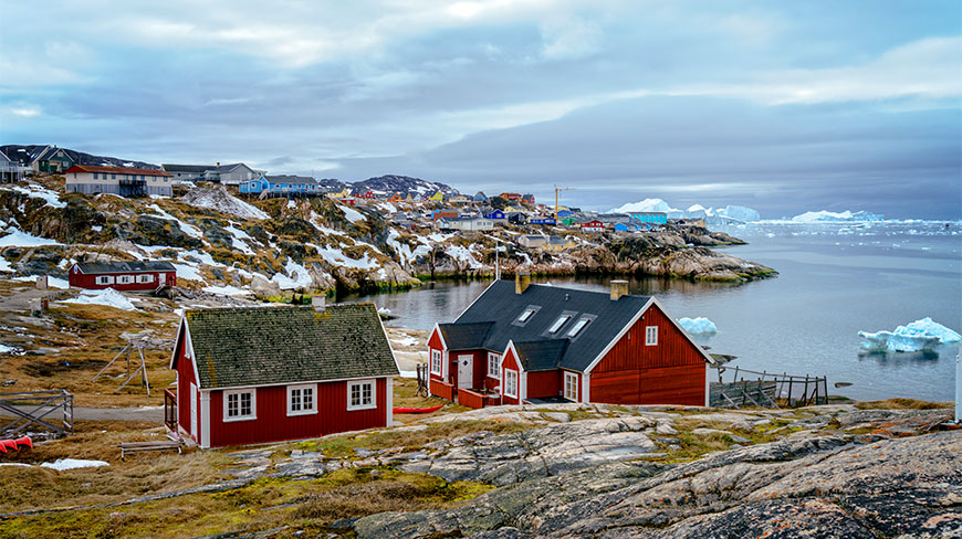Colorful red houses sit on the rocky, snow-dusted coastline of Disko Bay, with icebergs floating in the calm water under a cloudy sky.