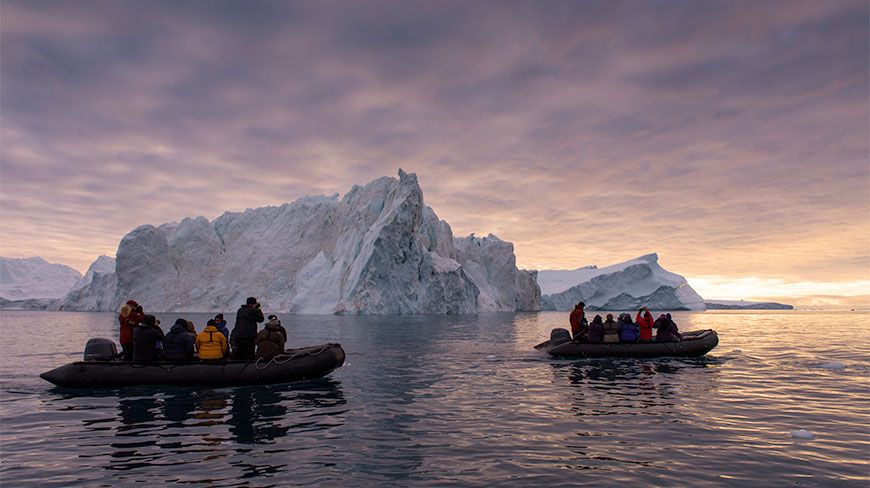 People in inflatable boats tour massive icebergs in Ilulissat, Greenland, under a colorful sunset sky.