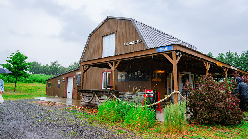 A rustic wooden Vinaigrerie building at a vinegar farm in Ile-d-Orleans, Quebec, featuring a large covered porch on a cloudy day.
