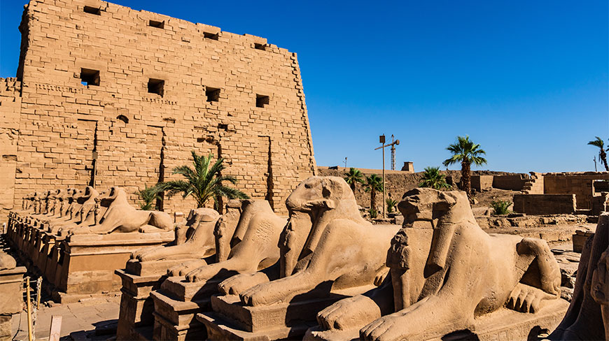 A row of ancient ram-headed sphinxes leads toward a massive stone pylon at the Karnak Temple complex in Luxor, Egypt.