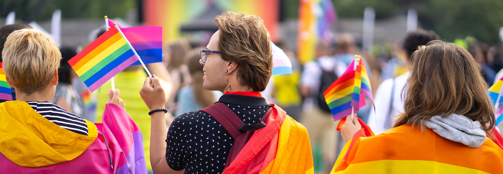 People celebrate at a pride parade, holding rainbow and bisexual flags while draped in colorful pride flag capes.