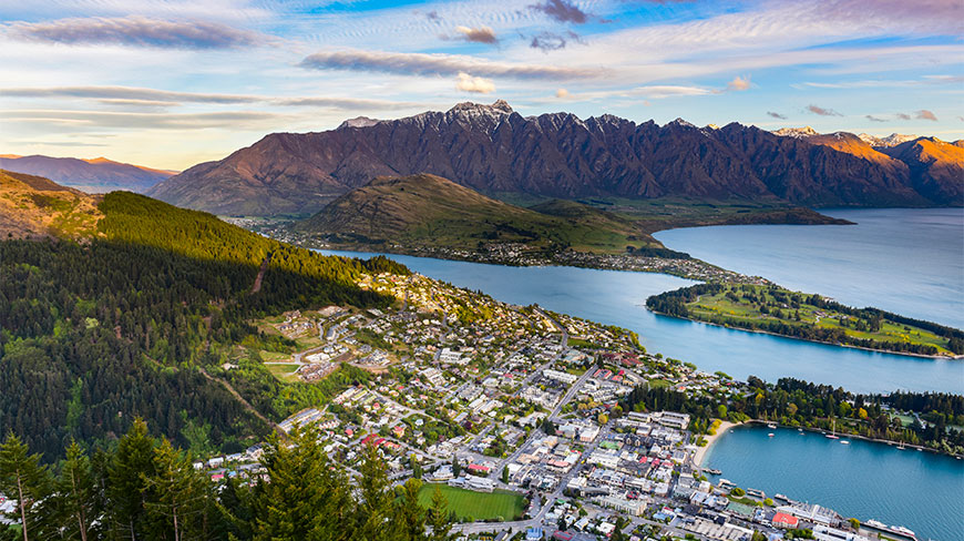 Aerial view of Queenstown, New Zealand, showcasing the town nestled along the blue lakefront with majestic mountain peaks rising in the background.