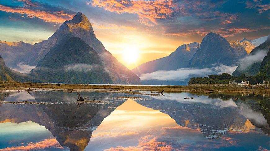 Vibrant sunset over Milford Sound in Fiordland National Park, New Zealand, with mountain peaks and golden clouds reflected in the still water below.