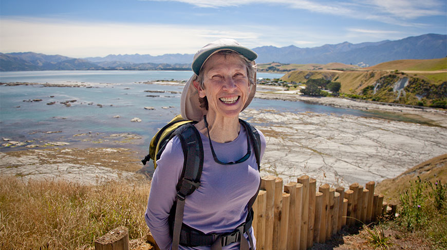 A smiling NZ participant in hiking gear stands outdoors overlooking a vast coastal landscape with rocky shores and distant mountains.