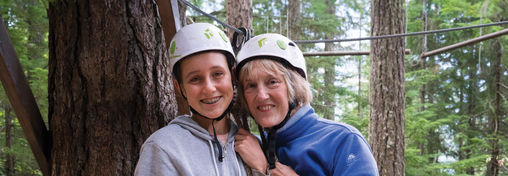 A grandmother and grandchild wearing helmets smile together during an outdoor adventure in a British Columbia forest.