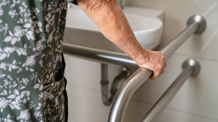 An older woman wearing a floral dress holds onto a metal handle bar for support in a bathroom next to a sink.